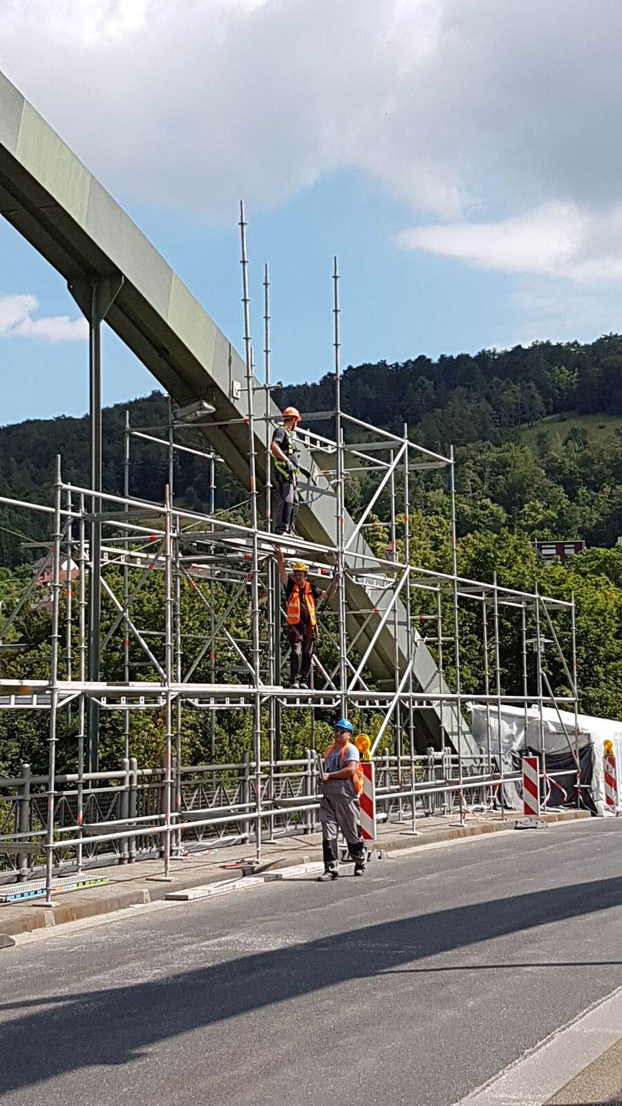 Eingerüstete Stahlbrücke über einer Straße, dahinter bewaldete Hügel